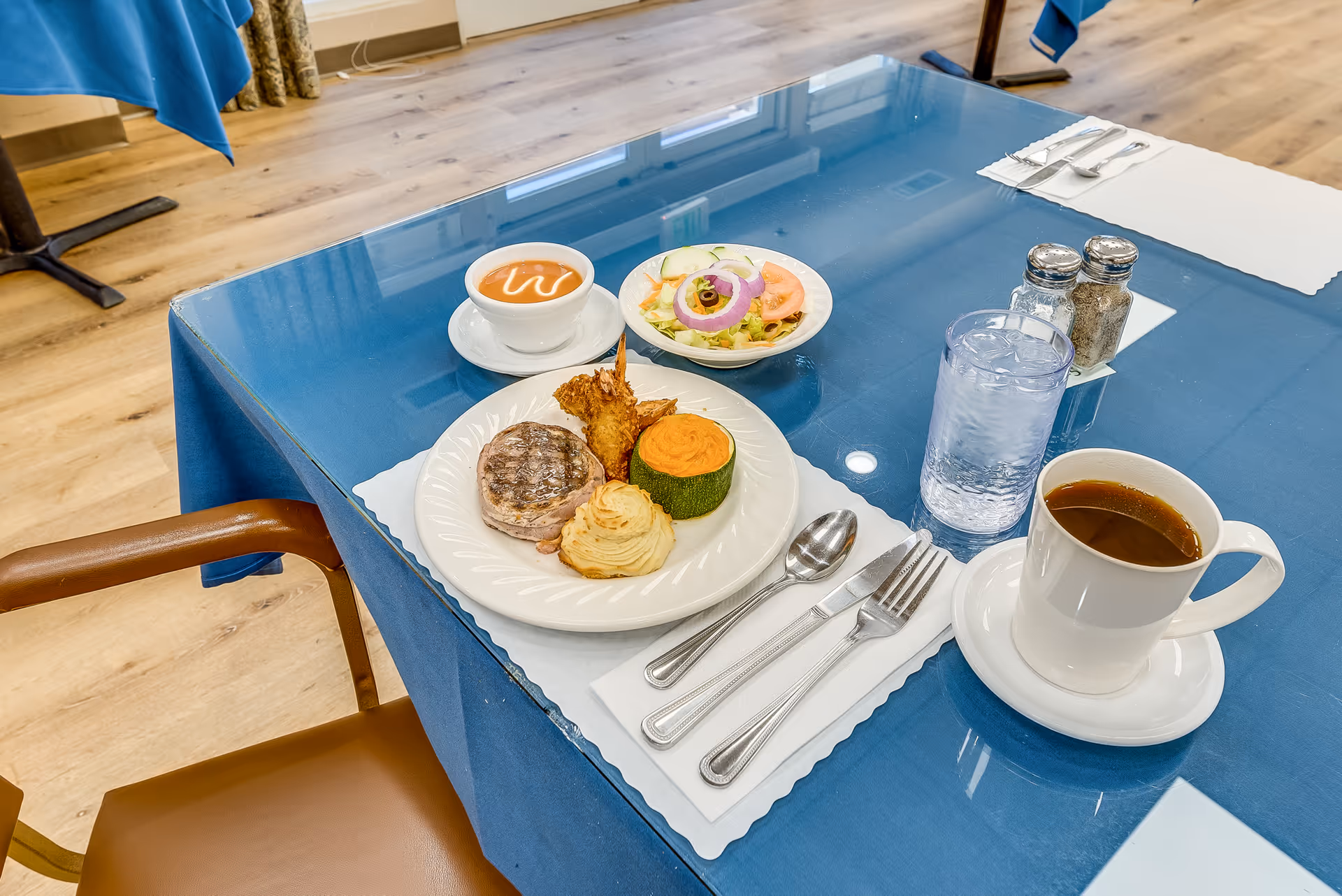 Table in a dining room set with a plated entree, salad, soup, a cup of coffee and a glass of water on a blue tablecloth.