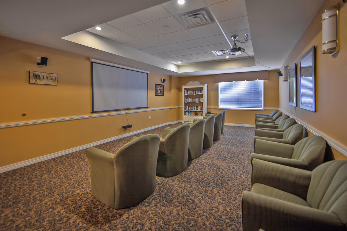 A cozy media room with two rows of green upholstered armchairs facing a large projector screen mounted on a mustard yellow wall. The room has patterned carpet, a bookshelf filled with books, a window with blinds, and framed pictures on the walls. Ceiling lights and a projector are visible.