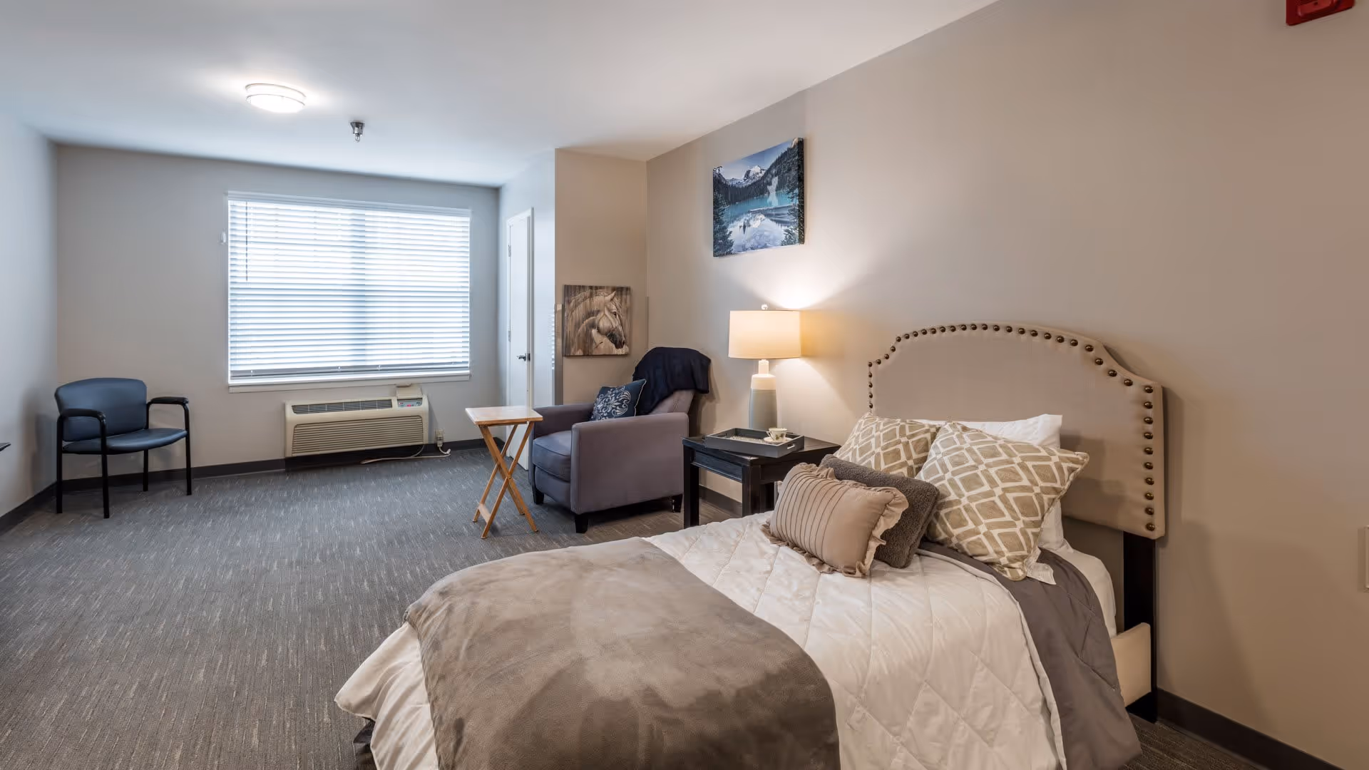 A cozy bedroom in a senior living facility with a neatly made bed featuring multiple pillows and a beige headboard. Next to the bed is a nightstand with a lamp and a small tray. A comfortable armchair with a pillow and a small wooden folding table are positioned near a window with blinds. The room has neutral-colored walls and carpeted flooring, with two framed pictures hanging on the walls.