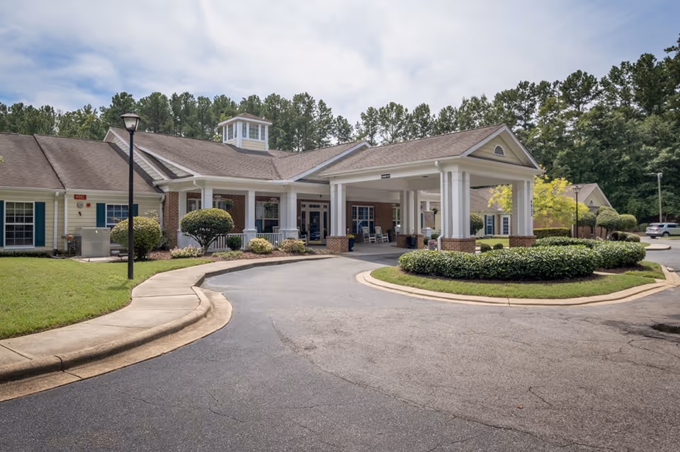 Exterior view of a single-story senior living facility building with a covered entrance driveway, surrounded by manicured bushes, trees, and a paved road under a partly cloudy sky.