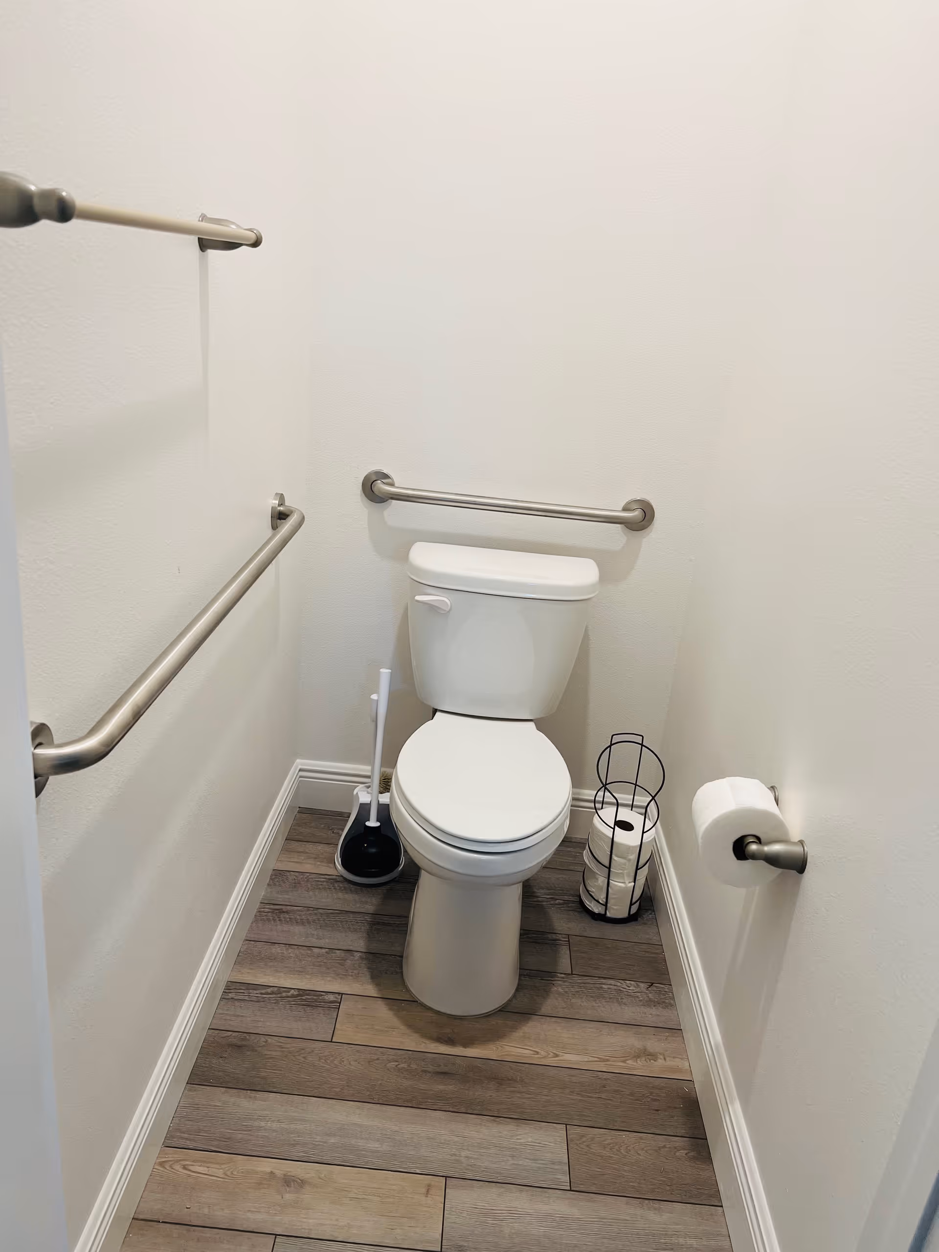 A small bathroom with a white toilet centered against the back wall. There are two metal grab bars on the left wall and one on the back wall behind the toilet. A toilet brush and holder are placed on the floor to the left of the toilet, and a black metal holder with three rolls of toilet paper is on the floor to the right. A toilet paper roll is mounted on the right wall. The floor has wood-like tiles and the walls are painted white.