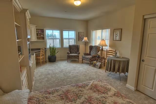 A cozy retirement facility room with a bed covered in a floral quilt in the foreground. The room has beige walls and carpet, two patterned armchairs with a small table and lamp between them near the windows, a round table with a tablecloth and a decorative item, a desk with a chair and built-in shelves, and a potted plant near the windows.