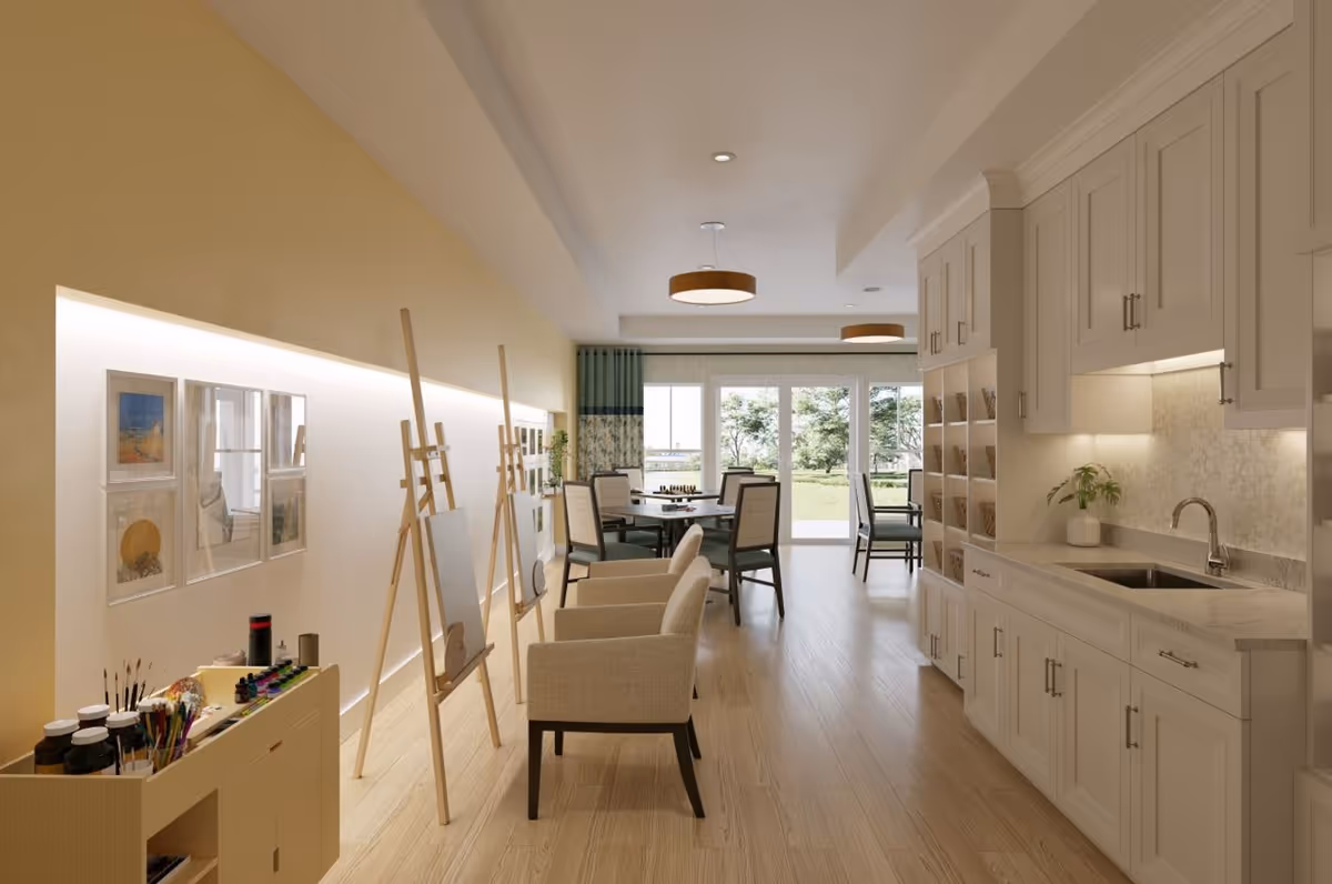 Bright multipurpose common room with easels and seating, a kitchenette on the right, and dining tables by large windows overlooking a lawn.