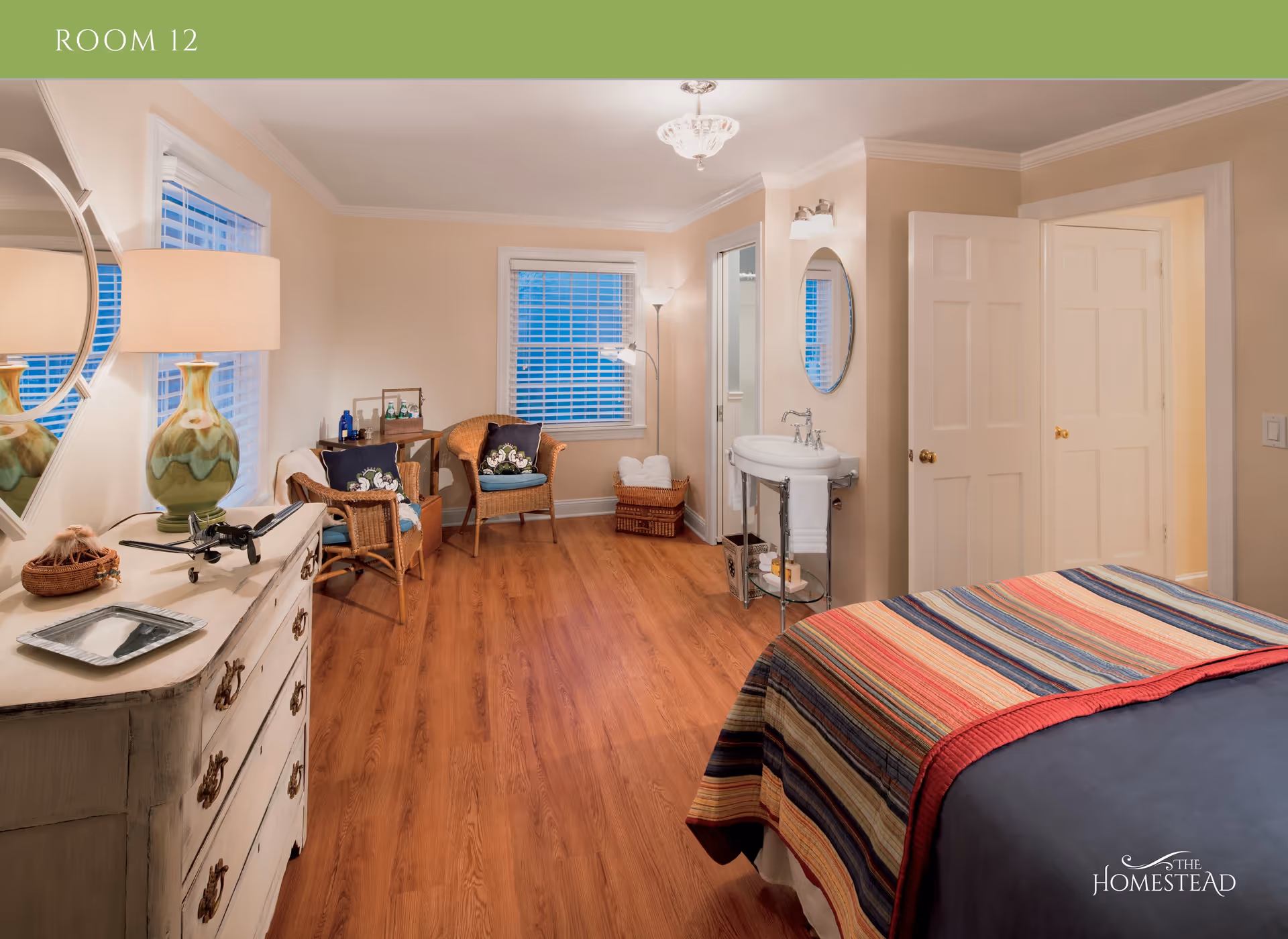 A cozy bedroom with wooden flooring, a bed covered with a colorful striped blanket, a white dresser with a green lamp and decorative items, two wicker chairs with cushions, a small table, a basket with towels, a wall-mounted sink with a round mirror above it, and two windows with white blinds.