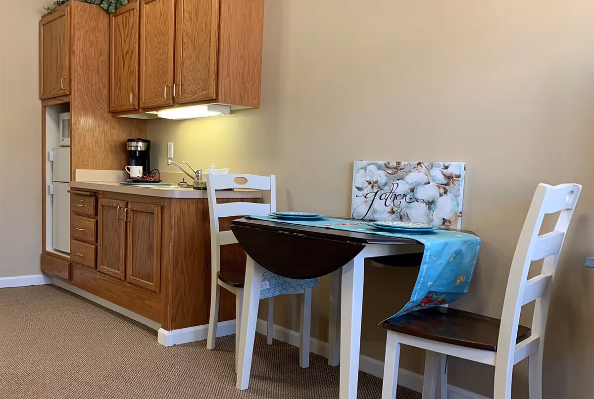 A small kitchen area with wooden cabinets, a countertop with a coffee maker, and a mini refrigerator. Next to the kitchen is a small dining table with a blue table runner, two plates, and two white chairs with dark wooden seats. A decorative sign with the word 'gather' and floral design is placed on the table against the wall.