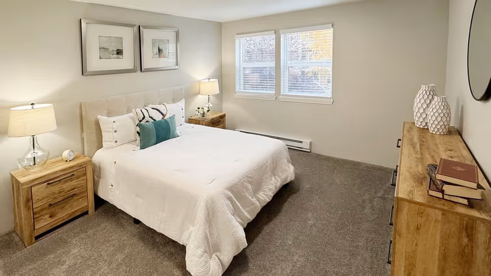 A neatly made bed with white bedding and decorative pillows in a bedroom. There are two wooden nightstands with lamps on either side of the bed. Two framed pictures hang above the bed. A wooden dresser with books and decorative vases is against the opposite wall. Two windows with blinds allow natural light into the room. The carpet is gray and the walls are painted light beige.
