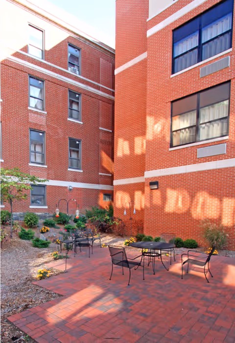 Outdoor patio area with red brick flooring and black metal table and chairs. The patio is surrounded by a garden with small bushes and flowers, and is enclosed by red brick building walls with multiple windows.