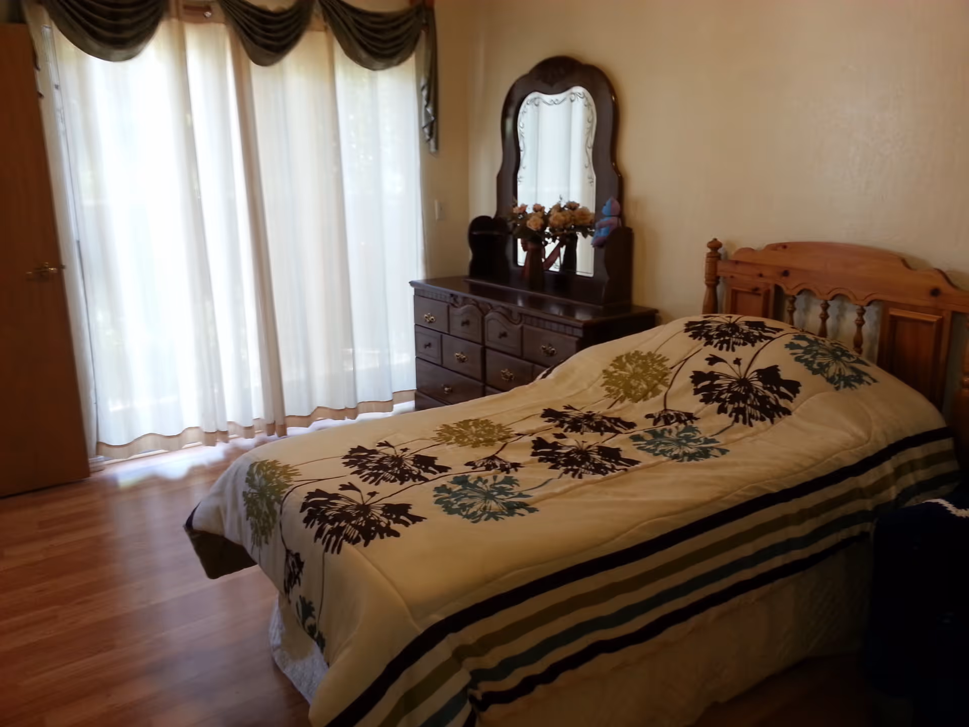 A bedroom with a wooden bed covered with a floral patterned comforter. Next to the bed is a wooden dresser with an ornate mirror and a vase of flowers on top. Sheer white curtains cover a large window or sliding door, allowing natural light to softly illuminate the room. The floor is wooden.