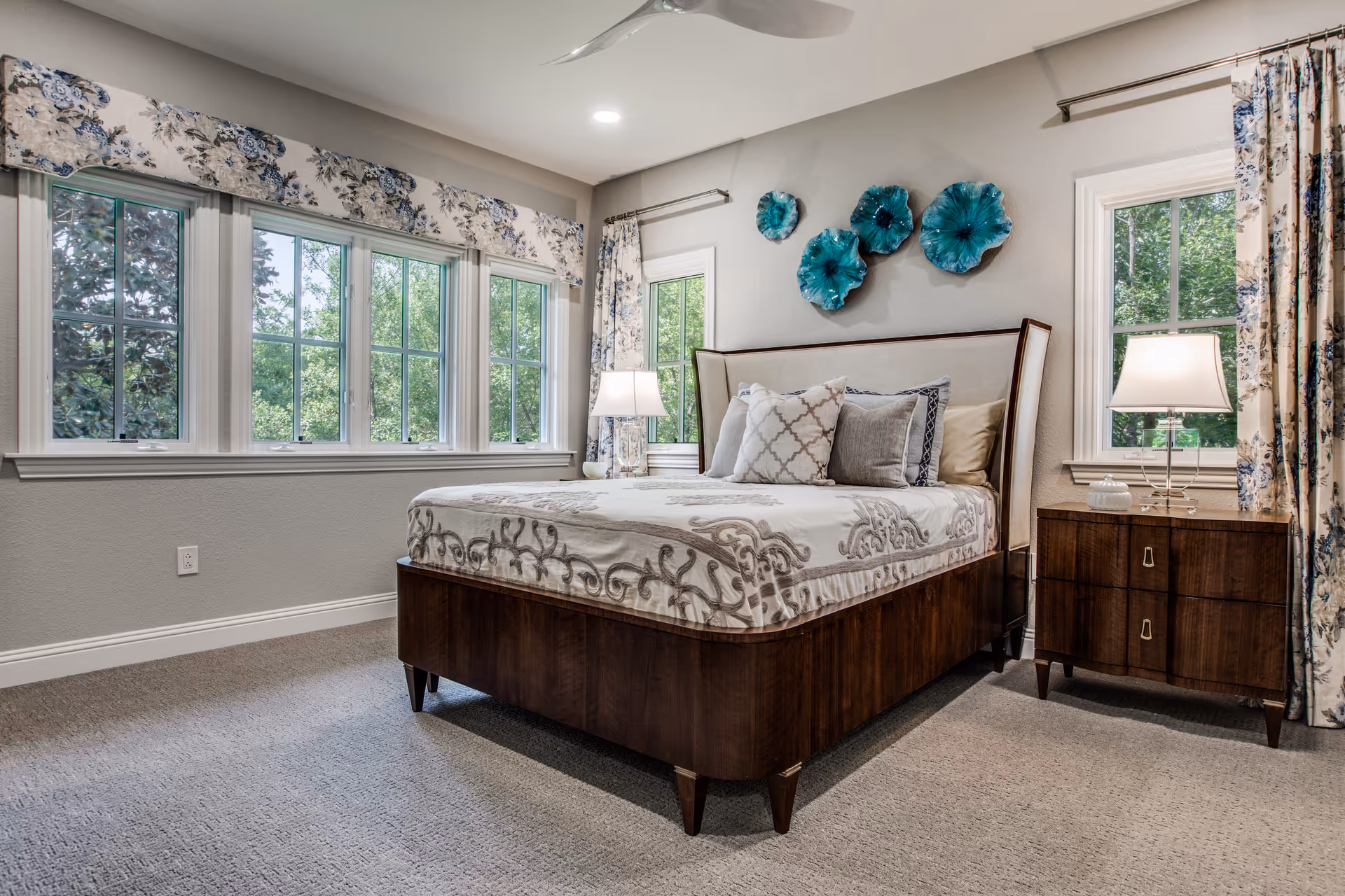 A well-lit bedroom with a large wooden bed featuring a patterned bedspread and multiple pillows. The room has three large windows with floral curtains and valances, two bedside tables with lamps, and decorative blue wall art above the bed. The carpeted floor and neutral wall colors create a cozy atmosphere.