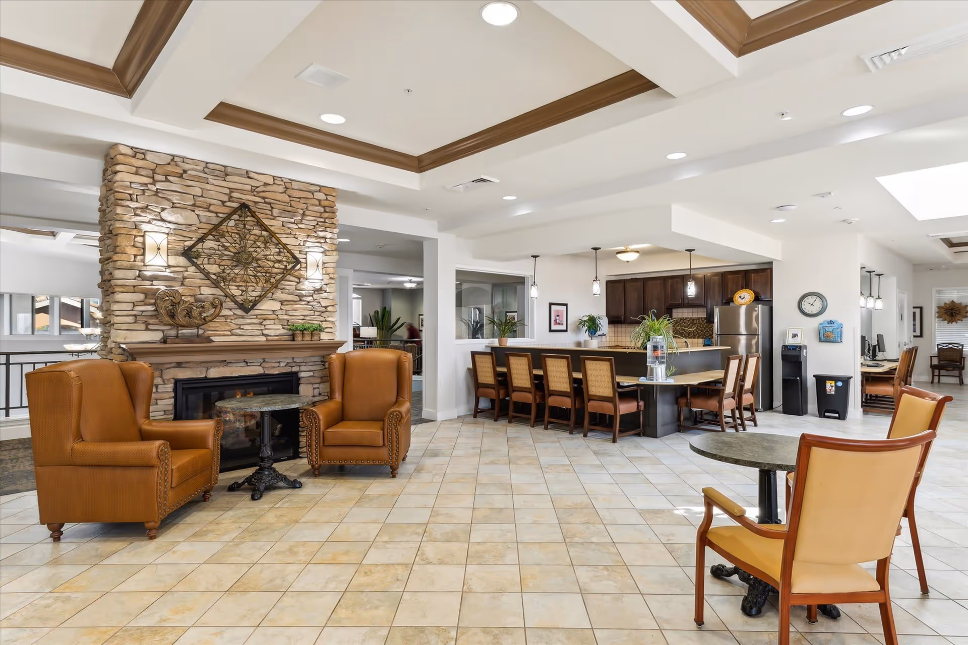 A spacious and well-lit common area in a senior living facility featuring a stone fireplace with decorative wall art above it, two brown leather armchairs with a small round table between them, a large kitchen island with several chairs, pendant lights hanging above, and a seating area with a round table and chairs in the foreground.