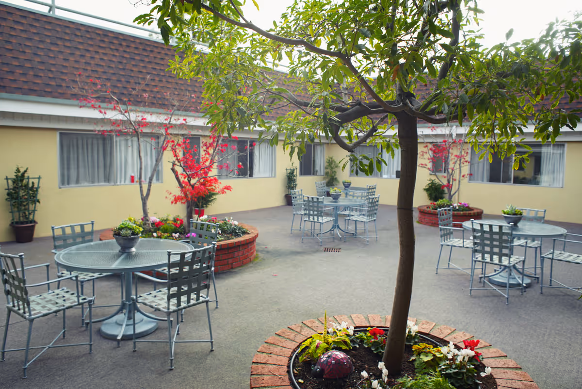 Outdoor courtyard area with several round metal tables and chairs arranged around planted trees and flower beds. The courtyard is surrounded by a single-story building with windows and beige walls.