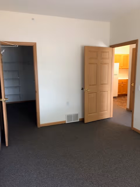 An empty room with beige walls and brown carpet. There are two open wooden doors; one door leads to a walk-in closet with shelves and a hanging rod, and the other door opens to a kitchen area with wooden cabinets and a white refrigerator.