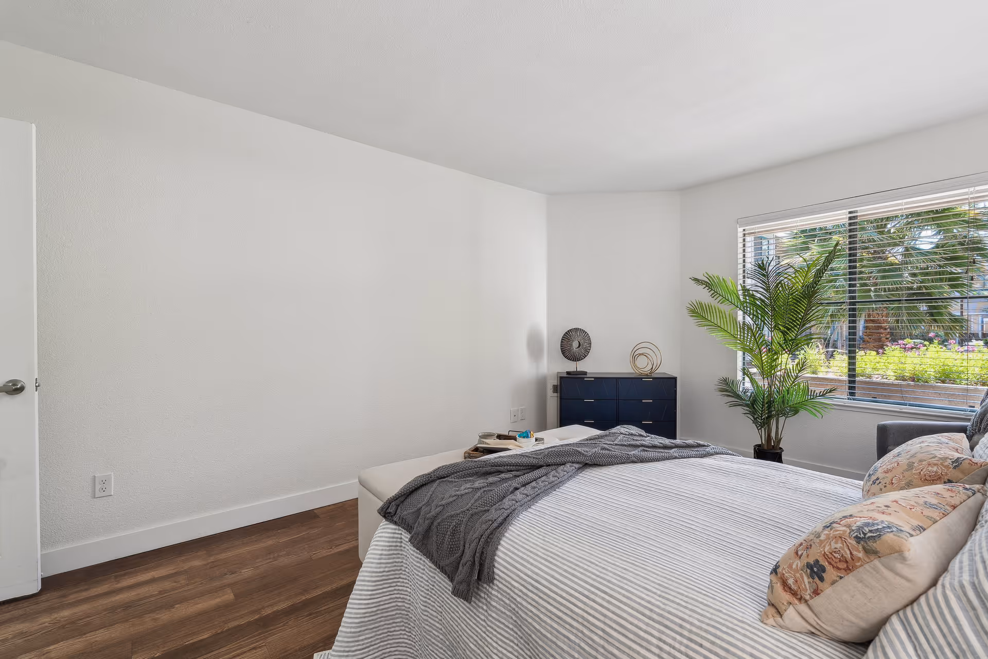 A bright bedroom with a large window showing outdoor greenery. The room features a bed with striped bedding and floral pillows, a gray throw blanket, a small dark blue dresser with decorative items on top, and a potted plant near the window. The floor is wooden, and the walls are white.