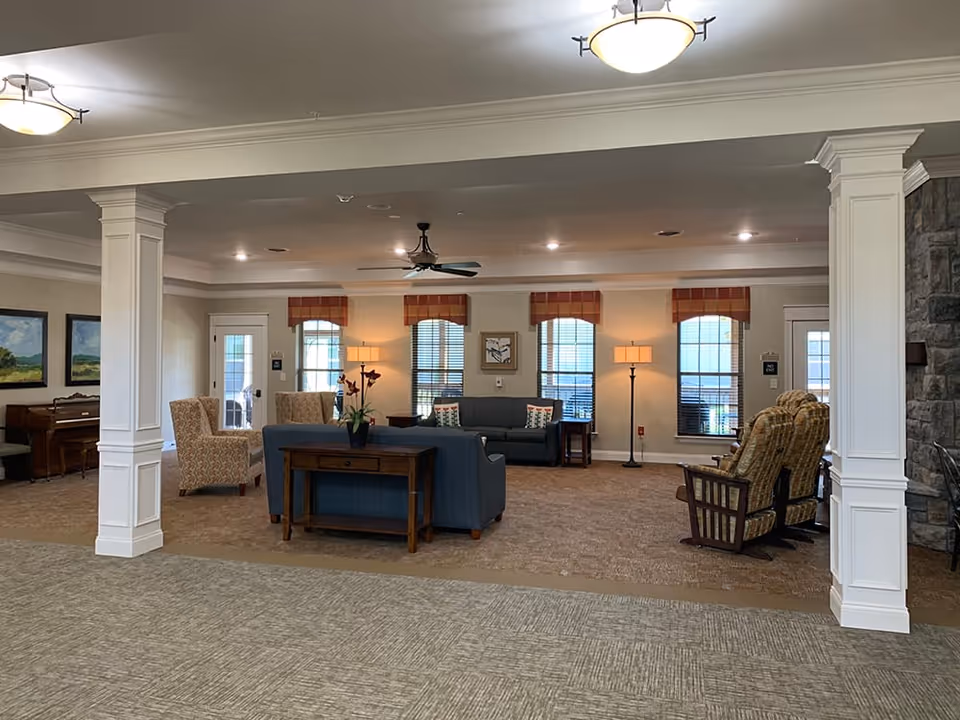 A cozy lobby area featuring comfortable seating, a piano, and large windows with natural light. The space is decorated with plants and artwork, creating a welcoming atmosphere.