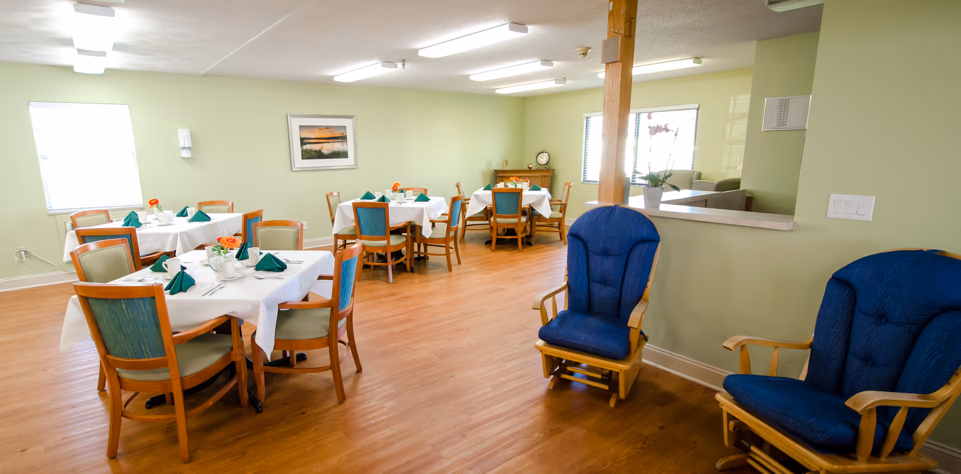 A bright dining room with several wooden tables covered with white tablecloths, each set with green folded napkins and small flower arrangements. The room has light green walls, wooden flooring, and two blue cushioned rocking chairs near a half wall with a window opening to another room.