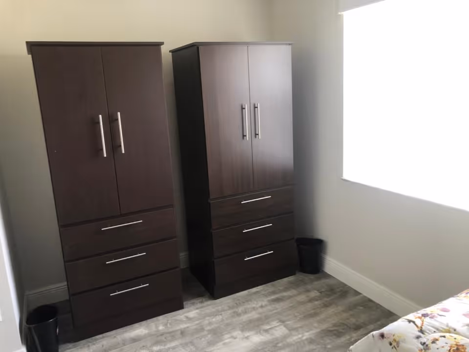 Two tall dark wood wardrobes with drawers in a bedroom corner beside a bright window and a partially visible bed.