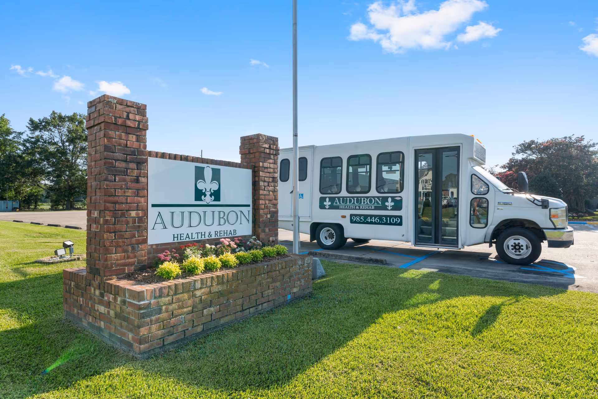 A brick sign for Audubon Health & Rehab with flowers planted at its base, situated on a grassy area next to a parking lot. Behind the sign, there is a white shuttle bus with the Audubon Health & Rehab logo and phone number parked in a handicapped parking space. The sky is clear and blue with a few clouds.