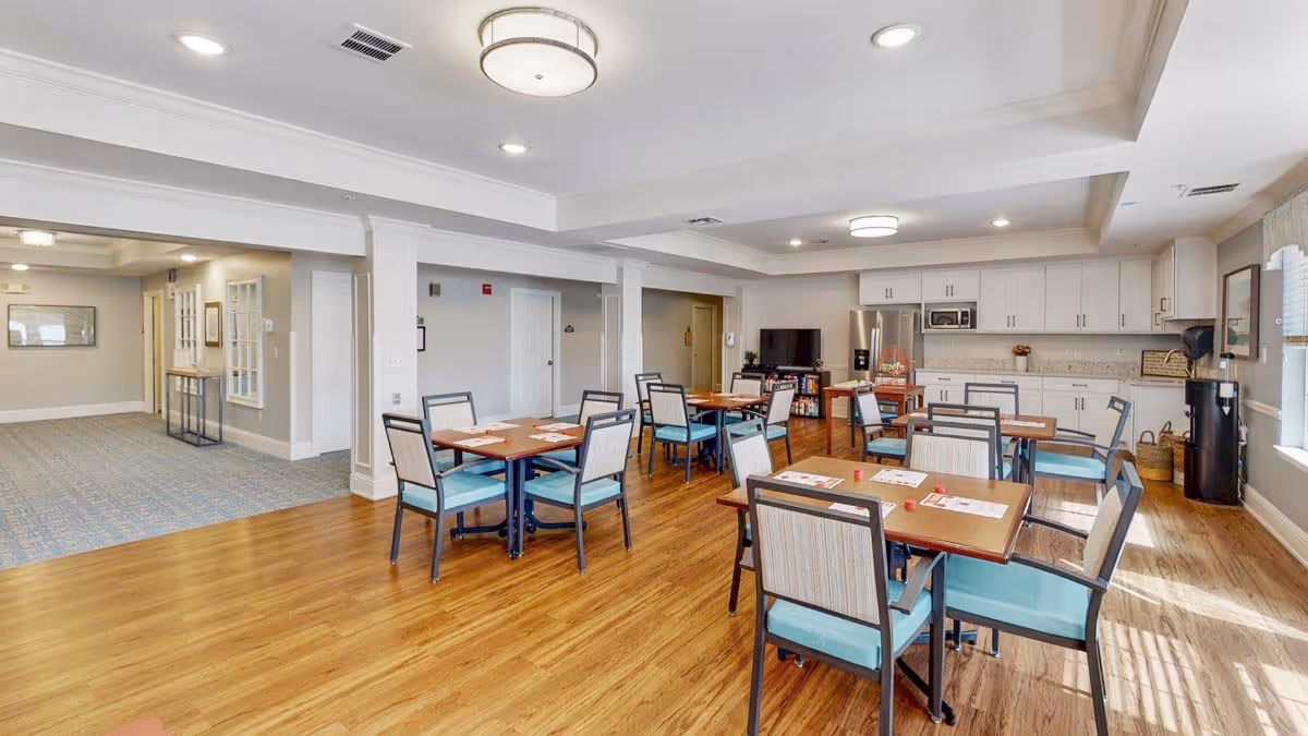 A bright and spacious dining room with several wooden tables and chairs with light blue cushions. The room features wood flooring, white walls, and a ceiling with recessed lighting and a central light fixture. In the background, there is a kitchenette area with white cabinets, a microwave, a refrigerator, and a coffee machine. Large windows on the right side allow natural light to fill the space.
