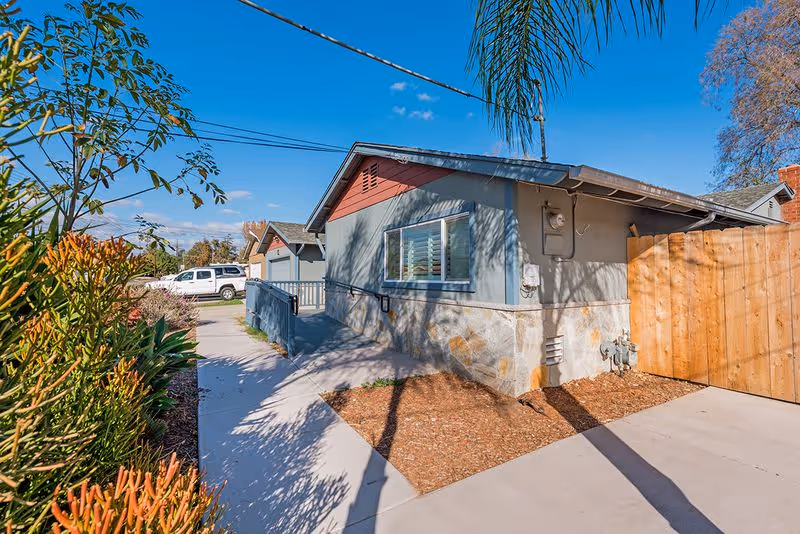 Exterior view of a single-story house with a concrete walkway, landscaped plants, a wooden gate, and a blue sky overhead.