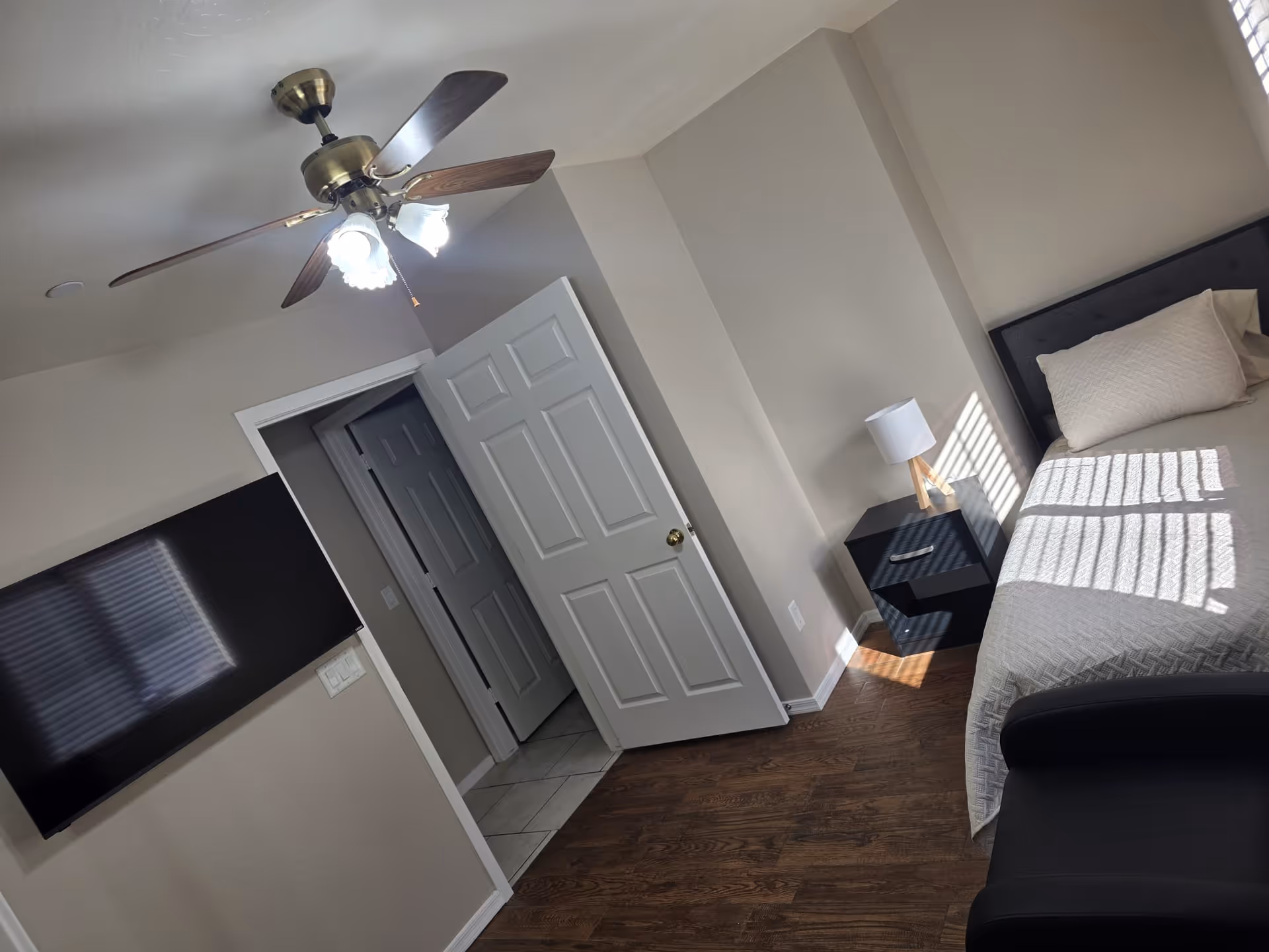 A bedroom with a bed covered in a light-colored quilt and a pillow, a black nightstand with a lamp, a ceiling fan with lights, a wall-mounted flat-screen TV, and an open door leading to another room with tiled flooring.