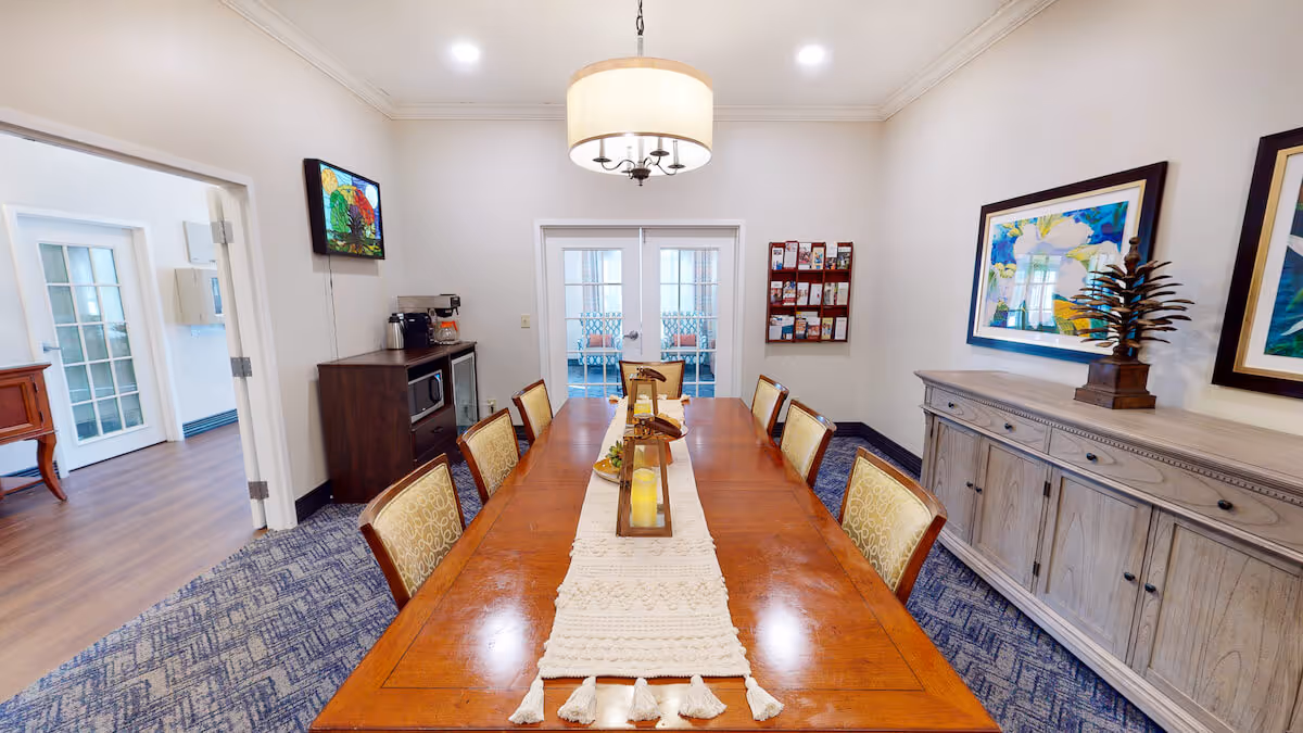A long wooden dining table with chairs, a chandelier, sideboard and artwork in a bright communal dining room.