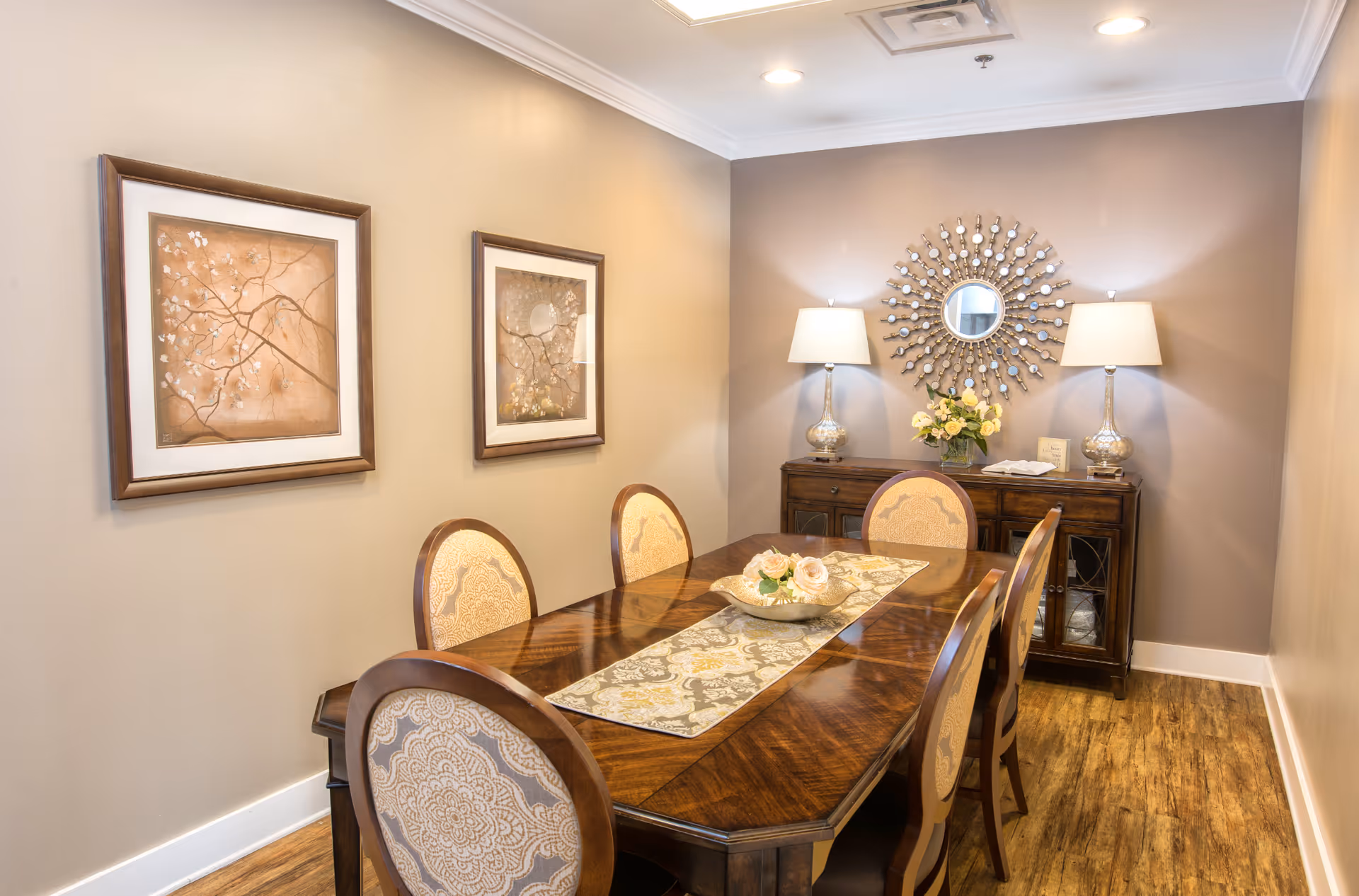 A warmly lit dining room with a wooden dining table and six upholstered chairs. The table is decorated with a patterned runner and a centerpiece bowl with flowers. On the back wall, there is a wooden sideboard with two matching lamps, a vase of flowers, and a decorative round mirror with a sunburst design. Two framed floral artworks hang on the left wall. The room has wood flooring and beige walls.