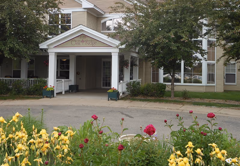 Entrance of Oak Ridge Assisted Living facility with a covered porch, surrounded by trees and flower beds with yellow and red flowers in the foreground.