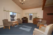Cozy lounge with blue carpet, armchairs, a piano, brick fireplace, and large windows.