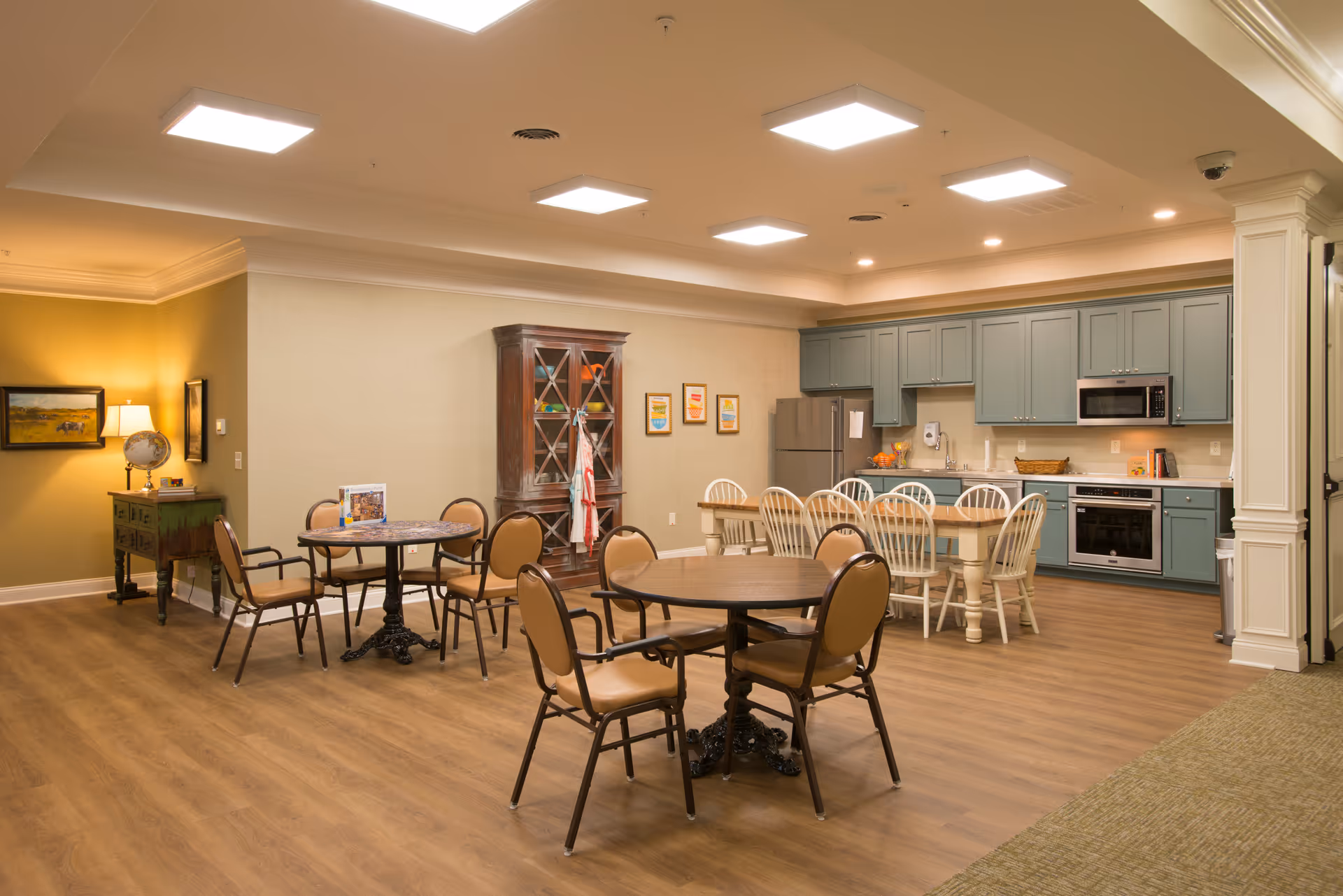 A spacious, well-lit dining area with multiple round tables and chairs. In the background, there is a kitchen with blue cabinets, a refrigerator, microwave, oven, and a long dining table with white chairs. The room has wooden flooring and warm lighting, with a small green side table and a globe lamp on the left side.