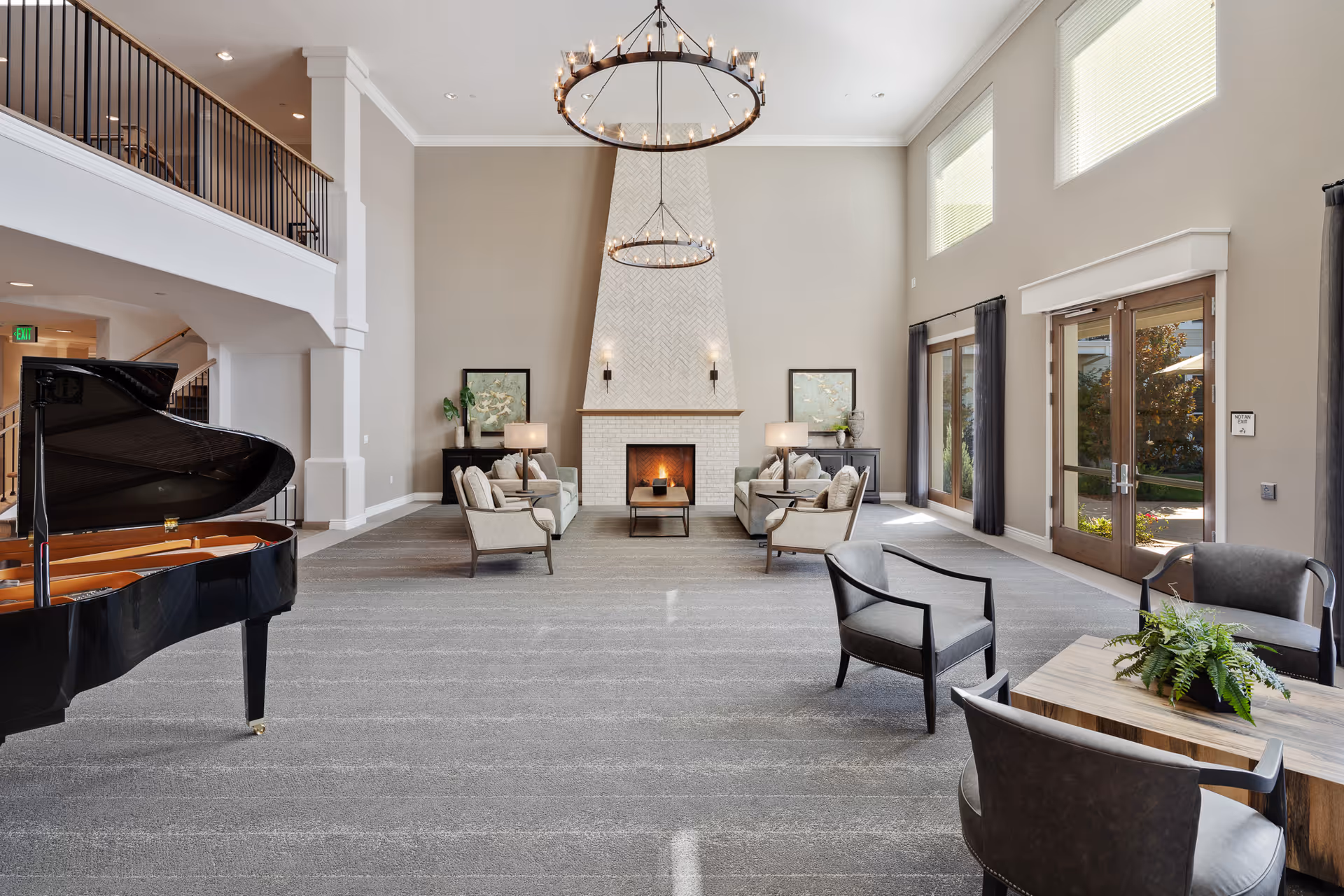 Spacious and elegant living room area with high ceilings, two large circular chandeliers, a white brick fireplace with a fire burning, and symmetrical seating arrangements including sofas and armchairs. A grand piano is positioned on the left side, and large windows and glass doors on the right let in natural light. The room has neutral tones with gray carpet and beige walls, and a small table with a plant is in the foreground.