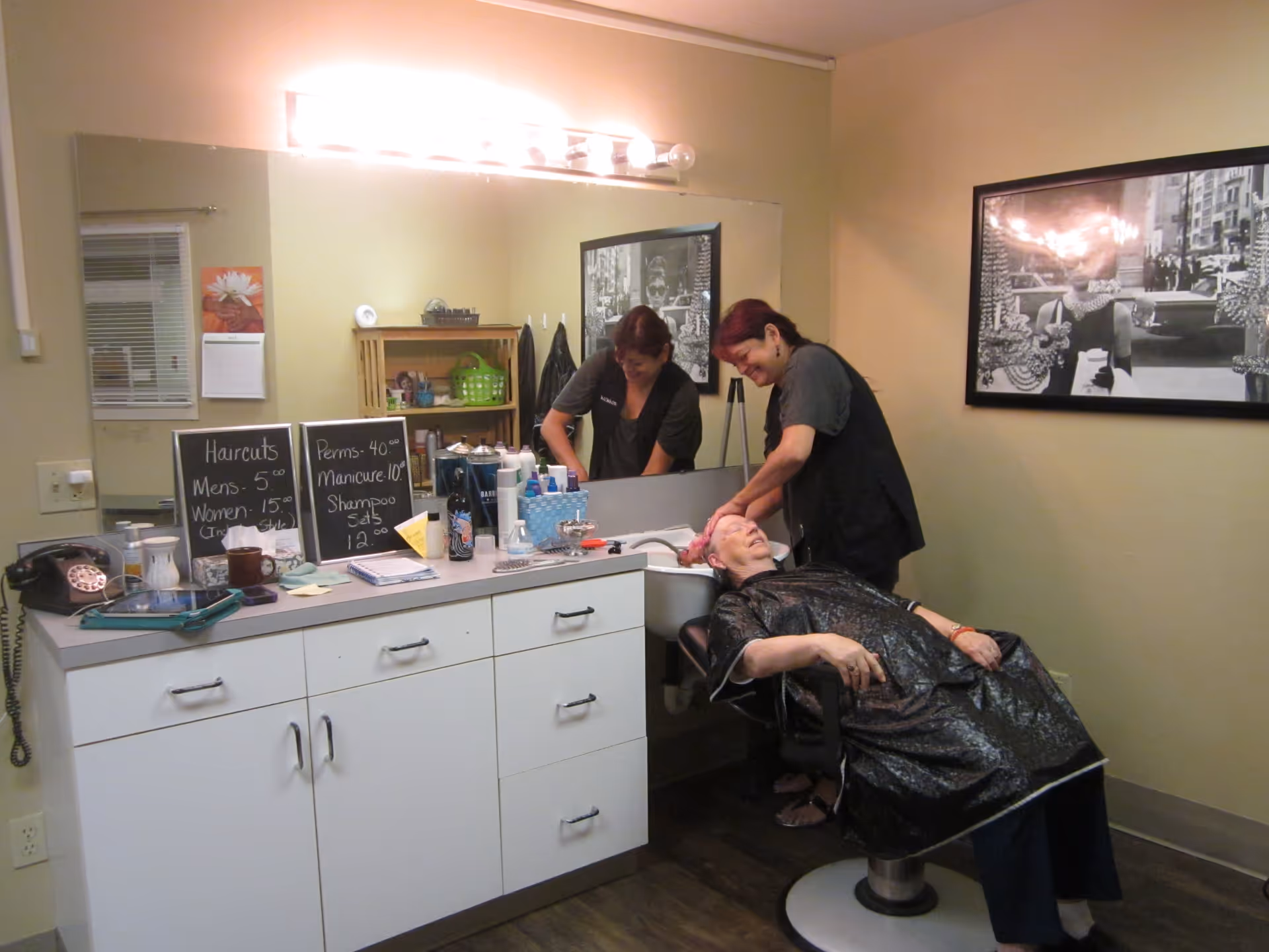 A hair salon area inside Sunnyside Assisted Living where a woman is washing an elderly woman's hair at a sink. The elderly woman is seated in a salon chair wearing a black cape. The counter has various hair products and two small chalkboards listing prices for haircuts, perms, manicures, and shampoo sets. A large mirror and a black-and-white framed photograph are on the walls.