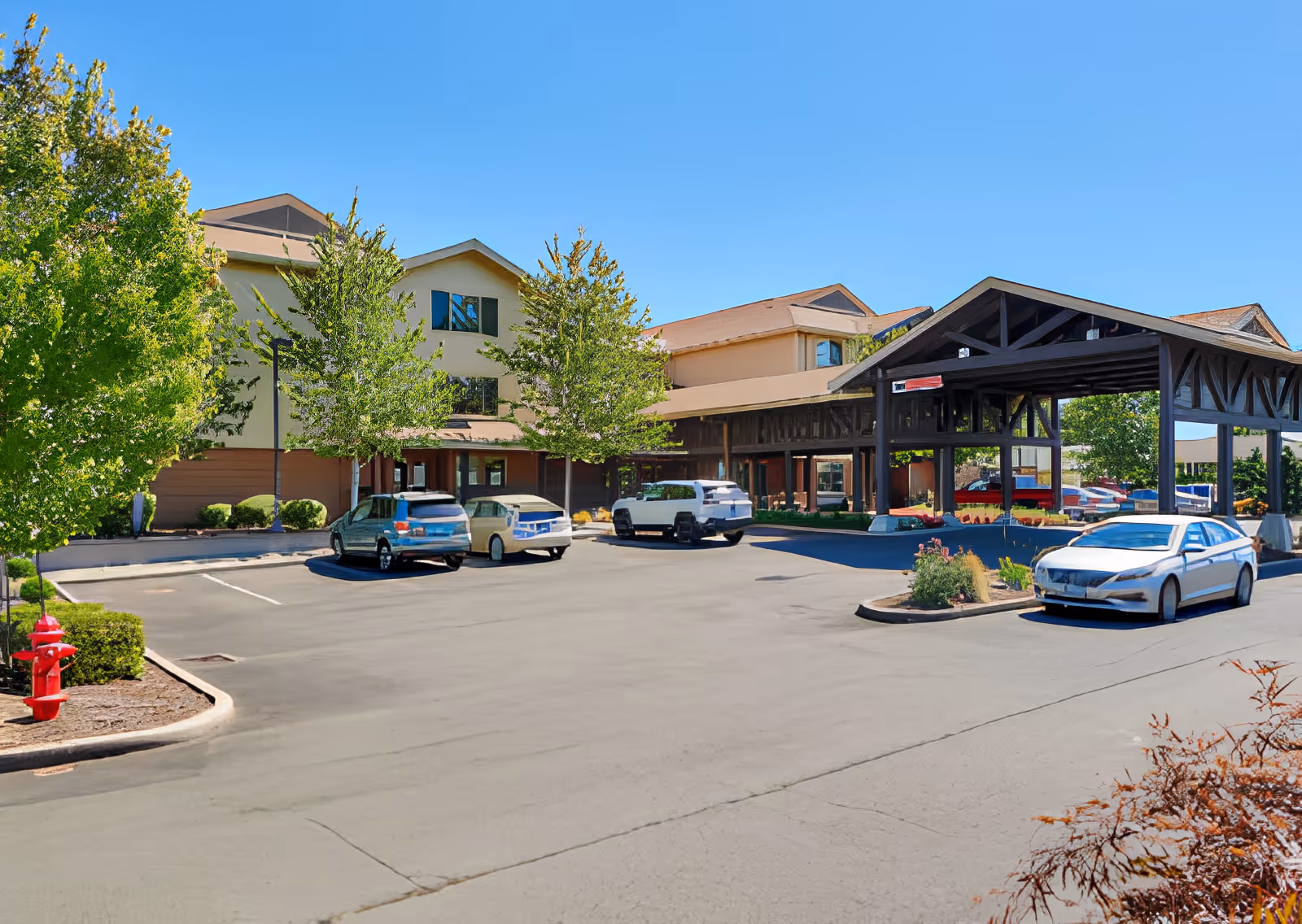 Exterior view of Elliott Residence senior living facility showing a parking lot with several cars, trees, and a covered entrance area under a clear blue sky.