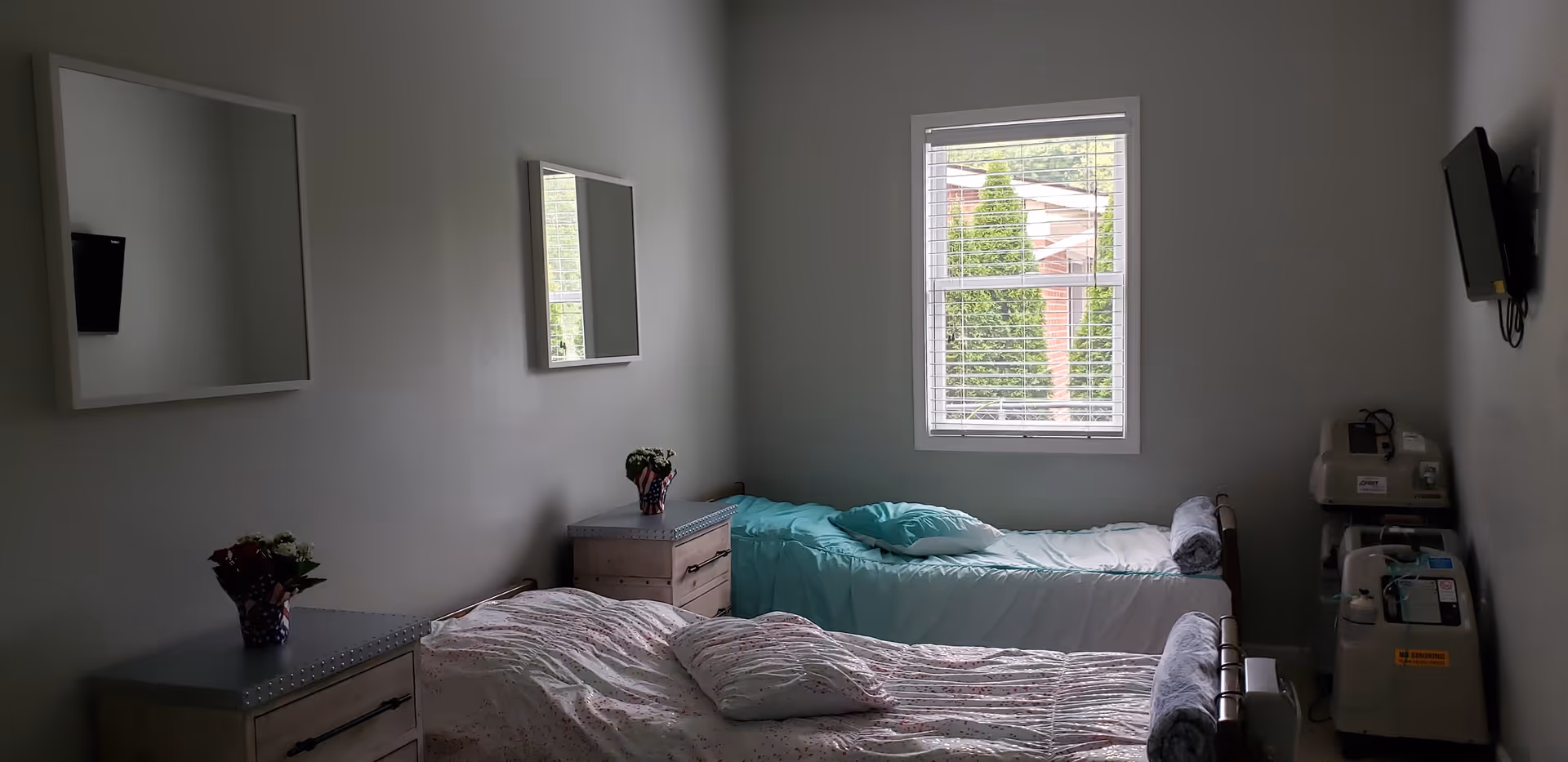 A small bedroom with two single beds placed parallel to each other. Each bed has pillows and blankets, with one bed covered in a light blue blanket and the other in a patterned white and pink blanket. There are two small bedside tables, each with a small flower arrangement. A window with blinds is centered on the far wall, letting in natural light. Two square mirrors hang on the left wall, and a wall-mounted TV is on the right wall above medical equipment.