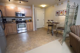 Small open kitchenette with stainless steel stove and refrigerator, wood cabinets, a round bistro dining table with chairs, and a decorative folding room divider.