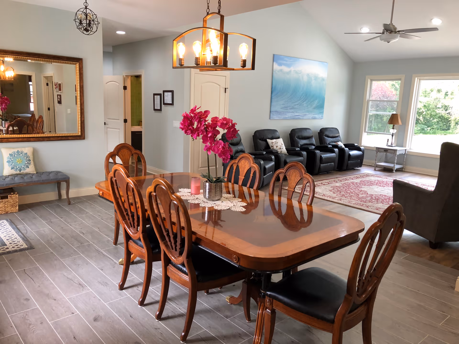 A spacious and well-lit common area with a wooden dining table and six chairs in the foreground. The table has a decorative centerpiece with pink flowers and a candle. In the background, there are four black recliner chairs arranged in a row facing a large window that lets in natural light. The room features light-colored walls, a large mirror on one wall, a ceiling fan, and a painting of a wave. The floor is covered with light wood-like tiles and a red patterned rug near the recliners.