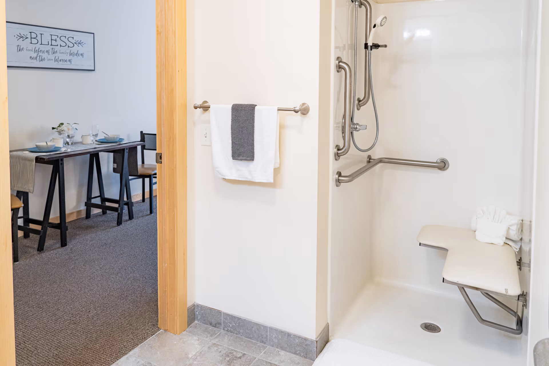 A bathroom shower area with a fold-down seat and multiple grab bars for accessibility. A towel rack with a white towel and a gray towel is mounted on the wall outside the shower. Through the open doorway, a dining area with a table set for a meal and chairs is visible. A wall decoration with the word 'BLESS' is hung above the dining table.
