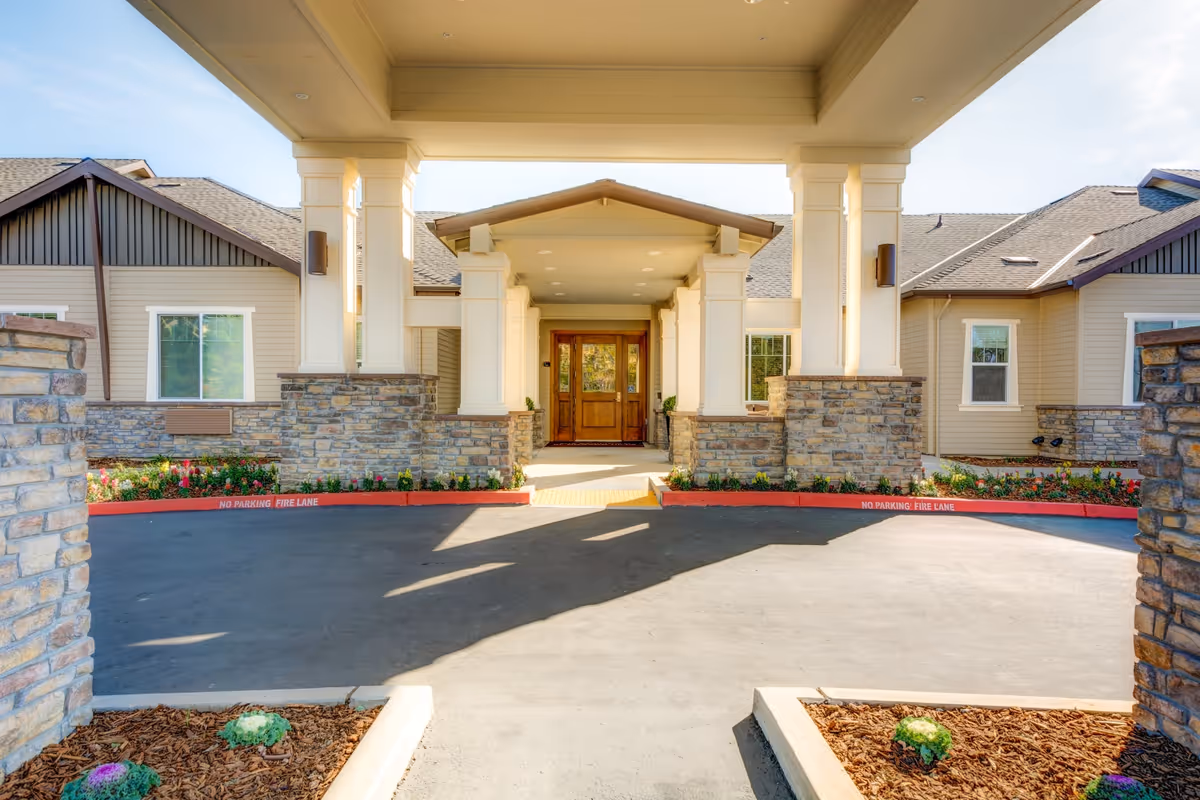 Covered porte-cochere entrance with stone columns, landscaping, and double wooden doors at a senior living facility.