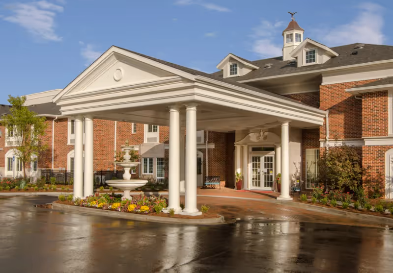 Entrance of a senior living facility named The Georgian Lakeside featuring a covered driveway with white columns, a decorative fountain surrounded by flowers, and a brick building with white trim under a blue sky.