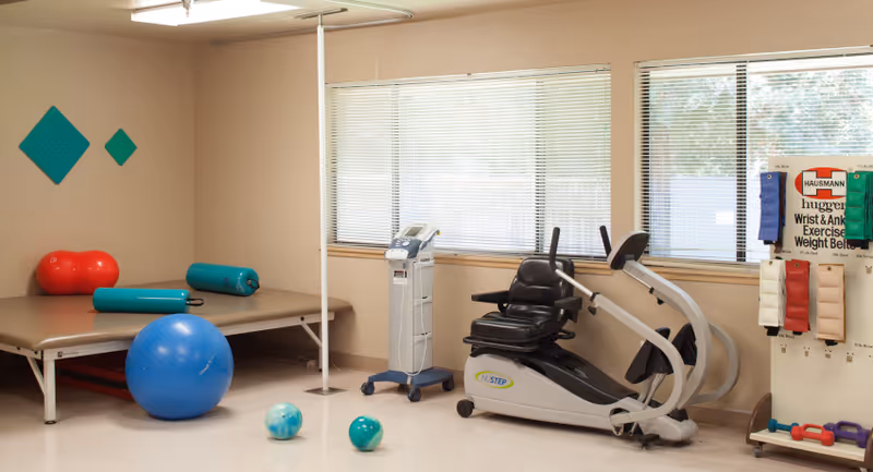 A physical therapy or exercise room with various equipment including a recumbent exercise bike, therapy balls, foam rollers, and wrist and ankle exercise weight belts displayed on a rack. The room has large windows with blinds and beige walls.