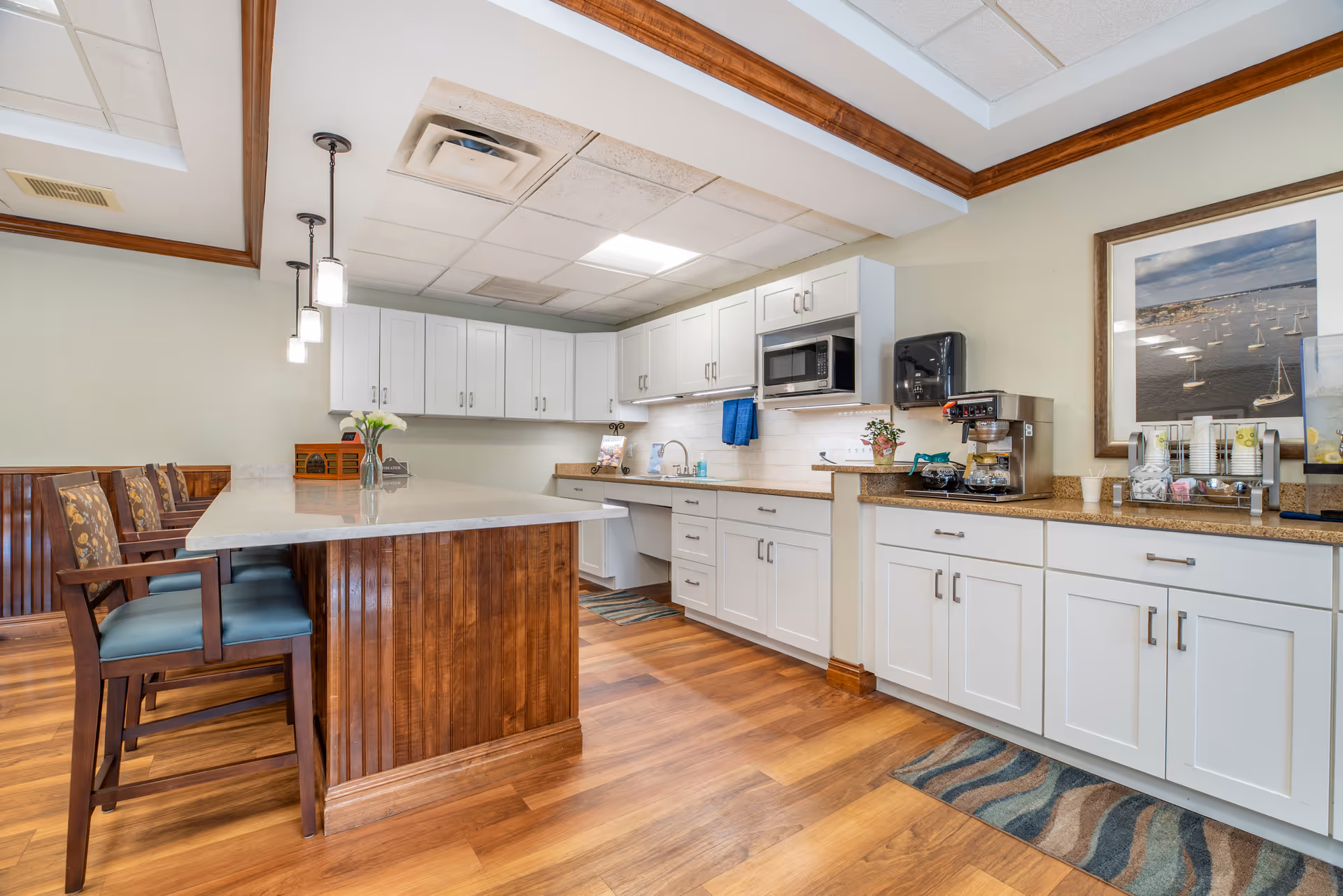 A bright kitchen area with white cabinets, a countertop with a coffee maker, microwave, and a sink. There is a wooden island with a marble top and chairs with blue cushions. The floor is wooden, and the ceiling has recessed lighting and pendant lights. A framed picture of sailboats on the water hangs on the wall.