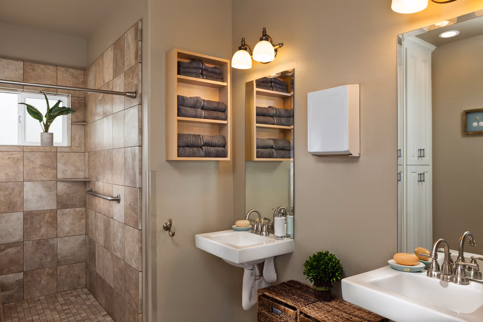 Tiled accessible bathroom with a walk-in shower, dual sinks, wall-mounted shelving stocked with towels, and decorative accents.