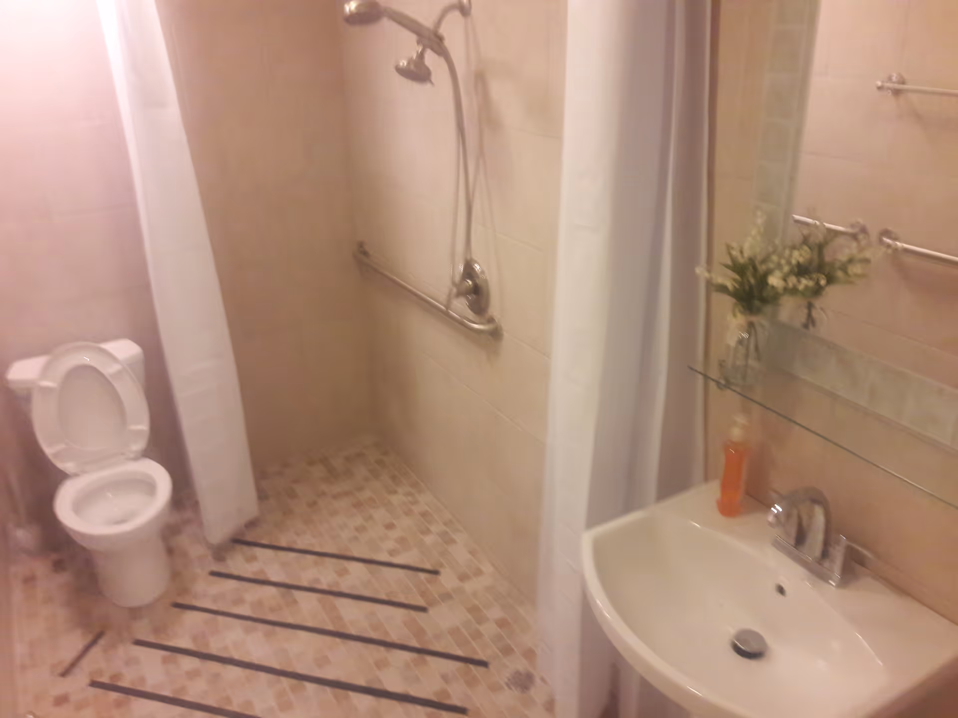 A bathroom with a toilet, a walk-in shower with a handheld showerhead and grab bar, a white shower curtain, a sink with a faucet, a mirror above the sink, a glass shelf holding a small vase with flowers and a bottle of soap, and tiled walls and floor with non-slip strips on the floor.