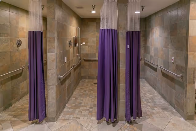 A tiled shower area with two shower stalls separated by purple and white shower curtains. Each stall has a wall-mounted showerhead, grab bars, and emergency pull cords. The walls and floor are covered with beige and brown tiles.