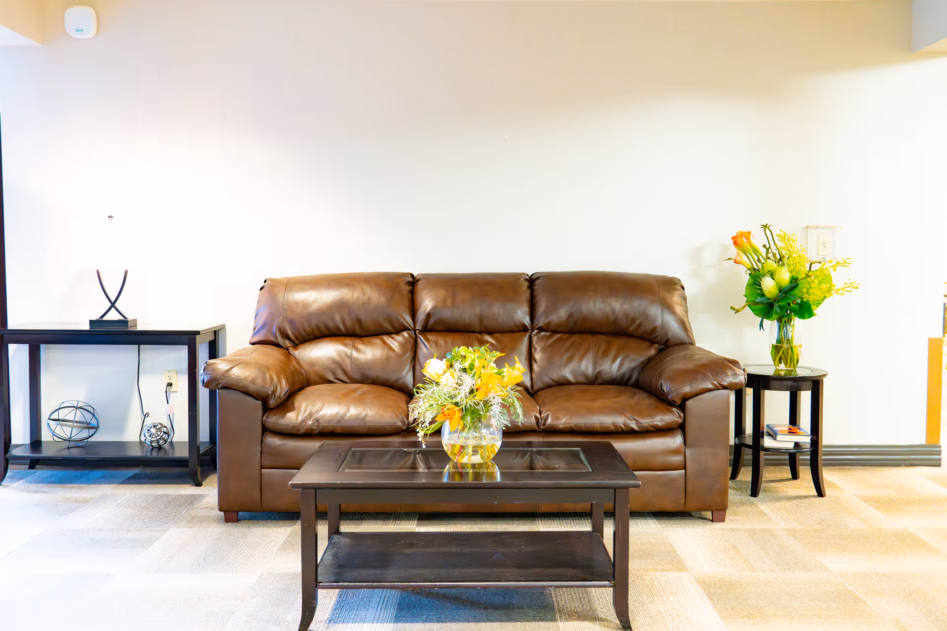 A cozy living room area with a brown leather sofa centered against a white wall. In front of the sofa is a dark wooden coffee table with a glass top, holding a vase of yellow and white flowers. To the left of the sofa is a side table with a modern lamp and decorative items, and to the right is a round side table with a vase of flowers and some books.