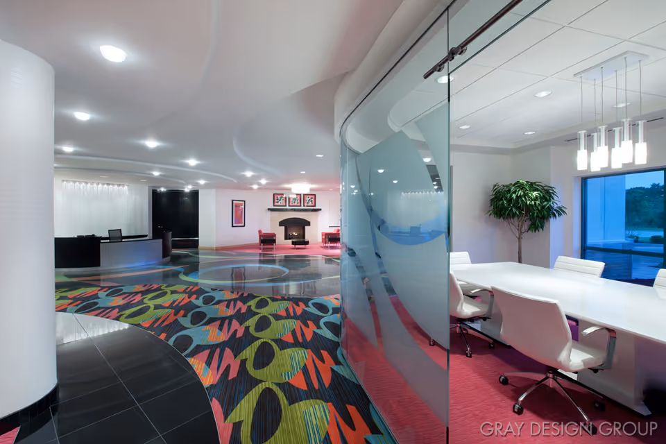 Modern interior space with a colorful patterned carpet, a curved white column, a reception desk area, and a glass-walled conference room with white chairs and a white table. There is a fireplace with framed artwork above it and a large plant near a window.