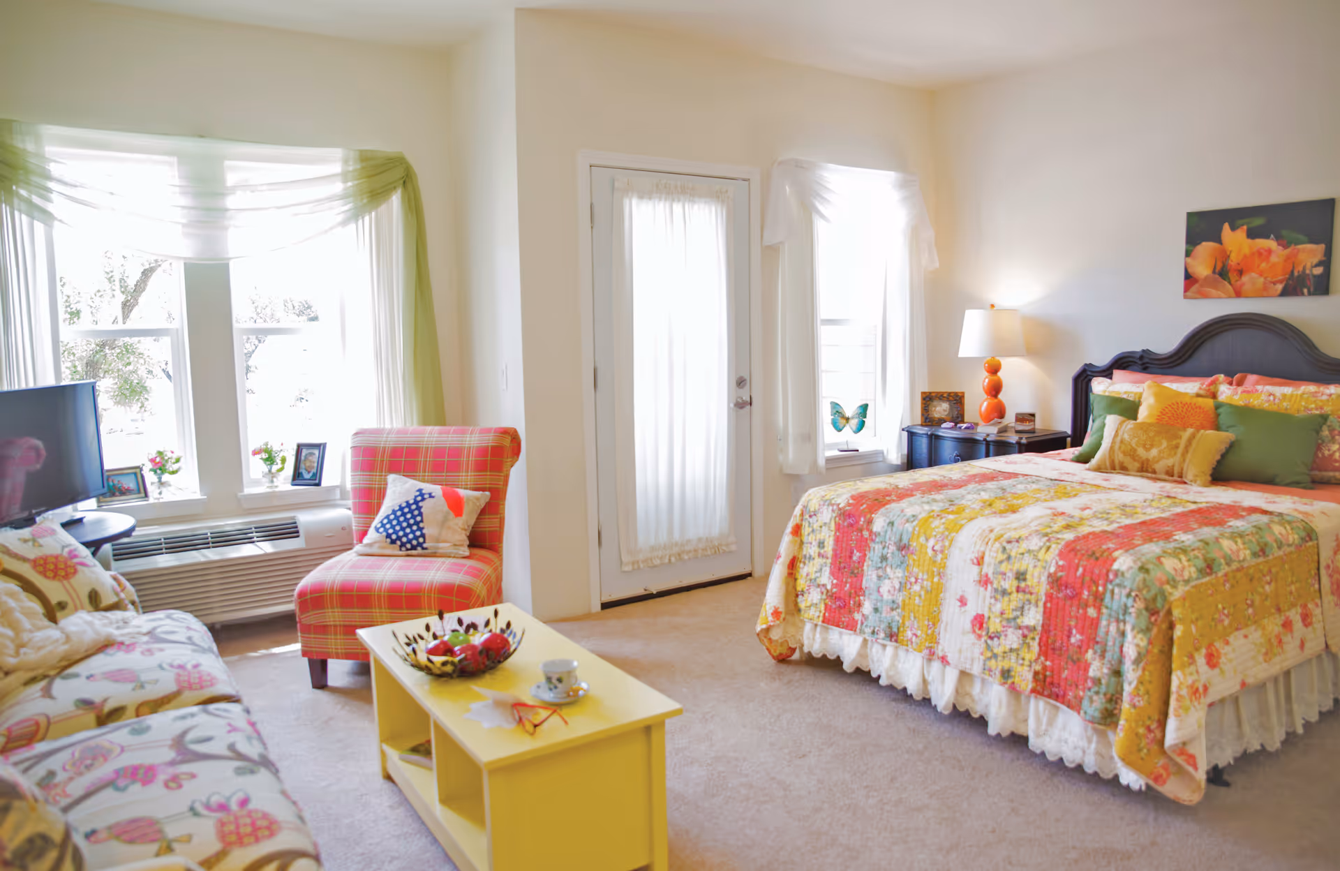 A bright and cozy bedroom featuring a large bed with a colorful floral quilt and multiple pillows. To the right of the bed is a dark wooden nightstand with a lamp and decorative items. A window with sheer white curtains is behind the nightstand. On the left side of the room, there is a plaid armchair with a decorative pillow, a small yellow coffee table with a bowl of fruit and a cup, and a floral-patterned sofa. Large windows with sheer green and white curtains let in natural light. A door with a sheer curtain is centered on the back wall.