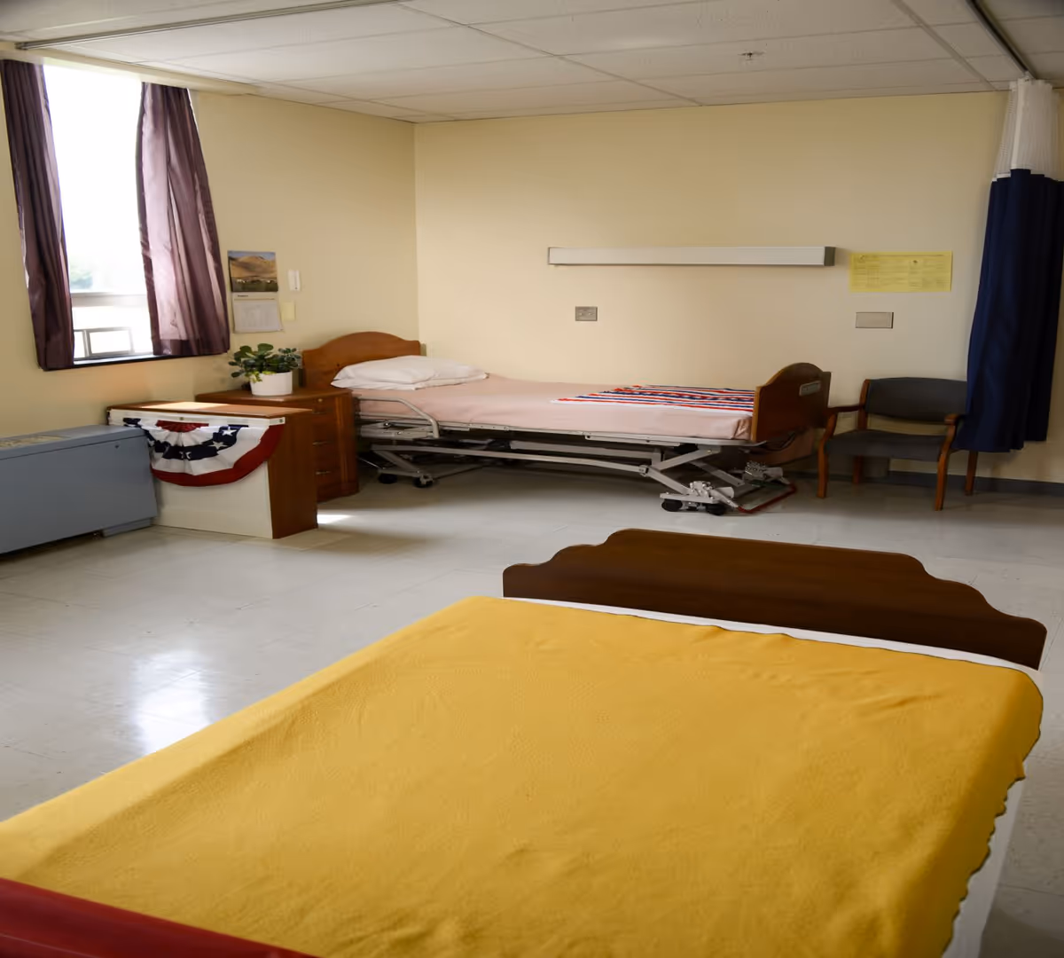 A simple room with two beds, one with a yellow blanket in the foreground and another hospital-style bed with a pink sheet and a patriotic red, white, and blue blanket in the background. The room has a window with purple curtains, a small wooden nightstand with a potted plant, a chair, and a privacy curtain on the right side.