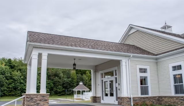 Entrance of a senior living facility with a covered drop-off area supported by white columns and stone bases. The building has beige siding with white trim and multiple windows. In the background, there is a small gazebo surrounded by greenery and trees under a cloudy sky.