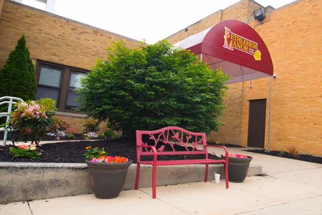 Outdoor entrance area of Bethlehem Manor Memory Care with a red bench, two large flower pots with colorful flowers, a green bush, and a maroon awning displaying the facility's name above a door on a brick building.