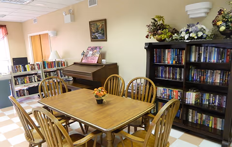 Wooden dining table with chairs in a communal room with bookshelves, a piano, and floral decorations.